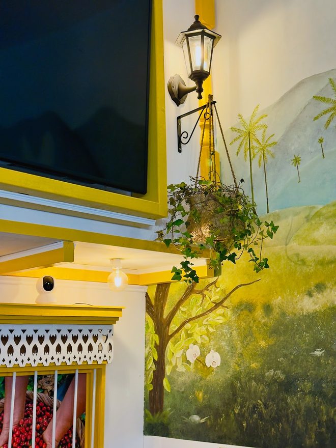 A yellow wall with a painted Colombian landscape mural, lantern and hanging plant